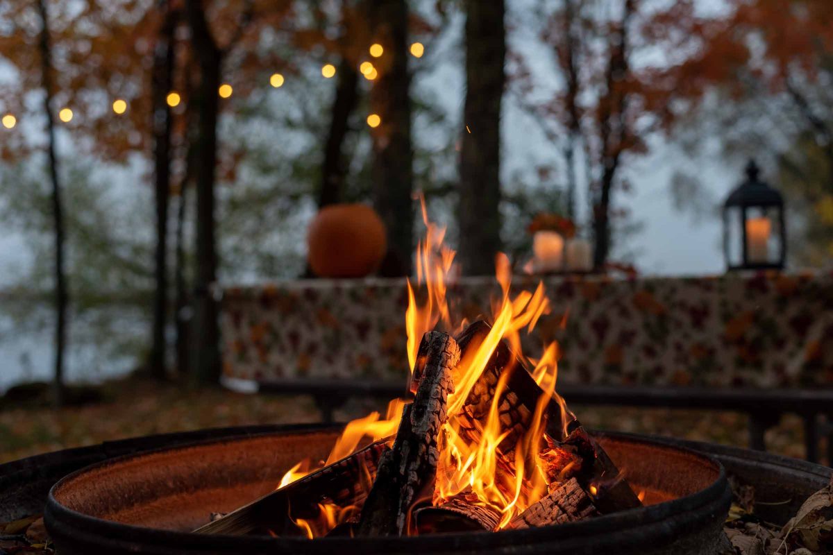 Flames dance in a round fire pit with twinkle lights and fall foliage in the background.