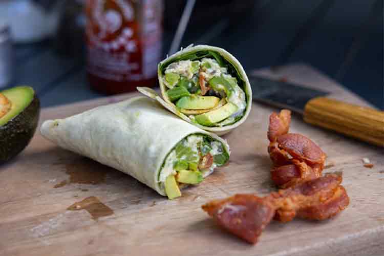 A camping breakfast burrito is cut in half on a cutting board with slices of bacon and avocado nearby