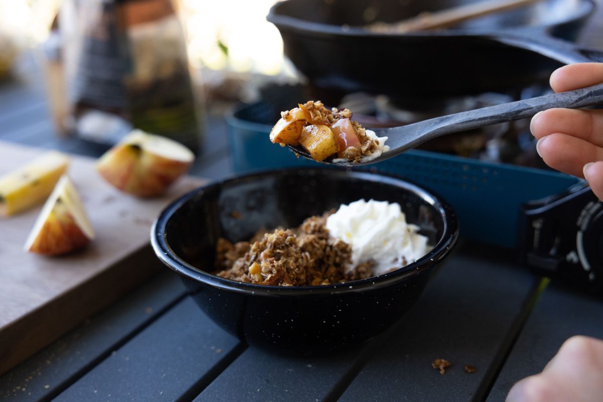 A hand holds a spoon over a black bowl filled with camp stove maple apple crisp and ice cream.