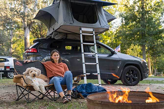 A camper and dog sit outside of a vehicle with a tent on top and a ladder leaning against the vehicle