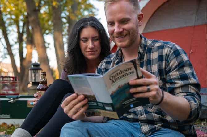 Two campers read through the Campspot Outdoor Almanac outside of their red tent.