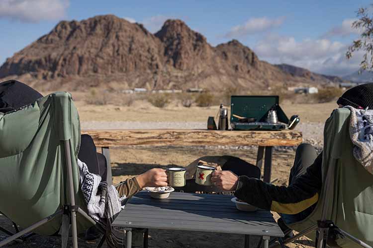 Two people sitting in front of camp site and rocky landscape drinking coffee.