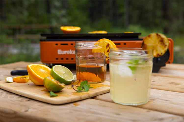 Two cocktails with lemon wedges and citrus cut up on a cutting board with a campstove in the background