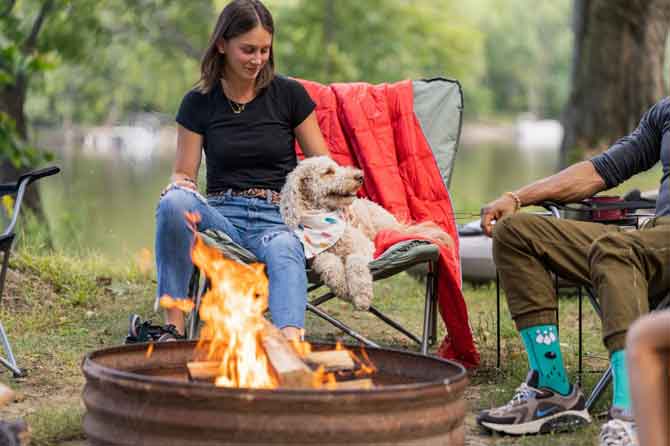 Woman sitting with dog in front of camp fire.